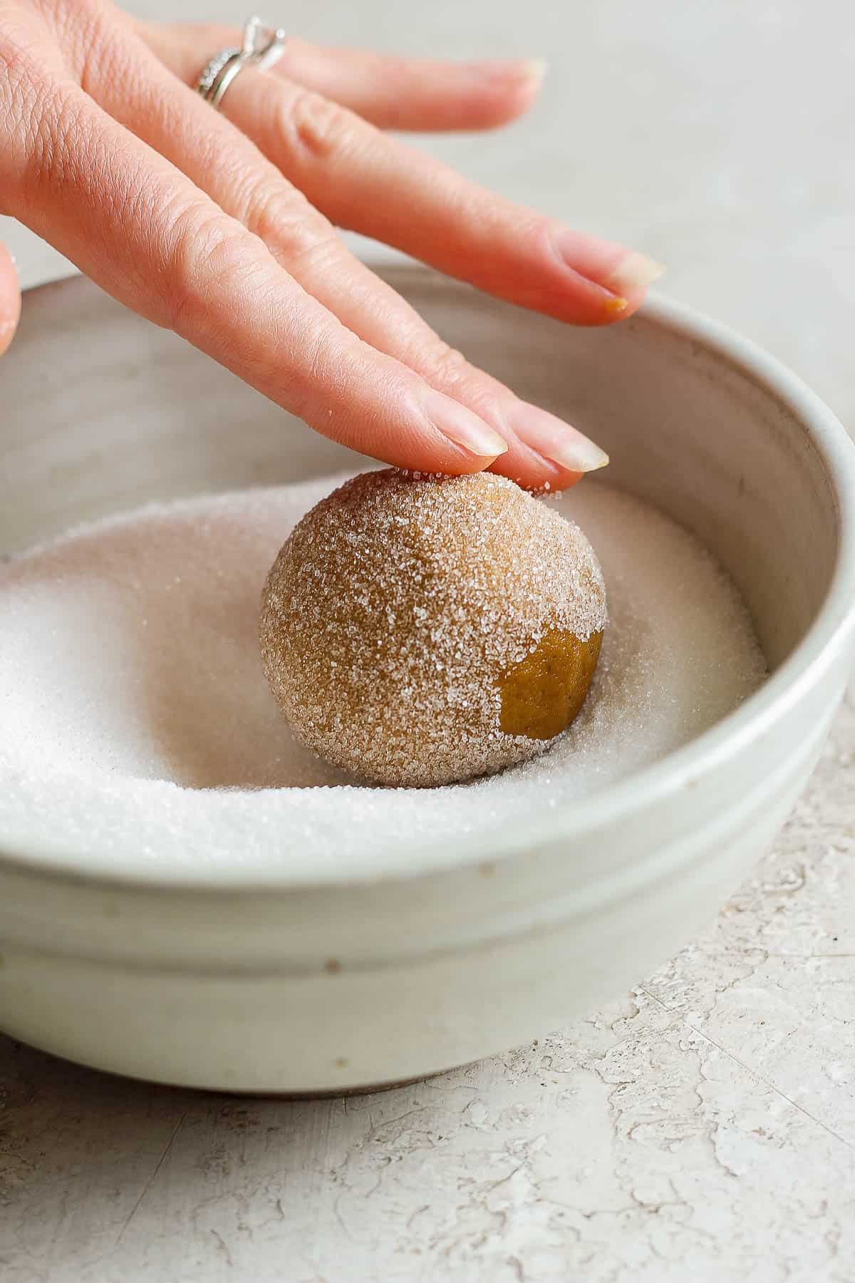 A ball of ginger molasses cookie being rolled in a bowl of white sugar.