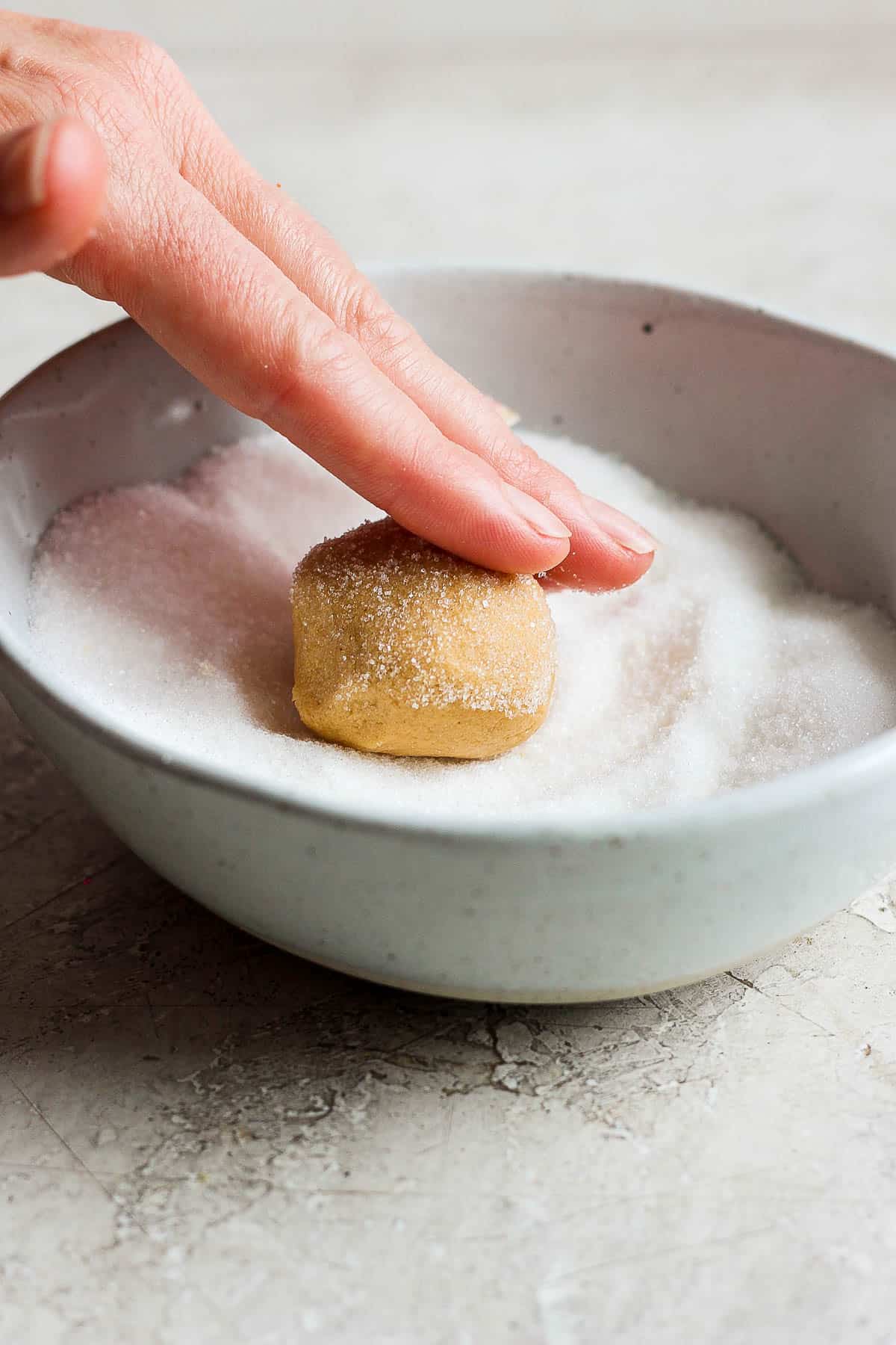 A hand rolling one dough ball in a bowl of sugar.