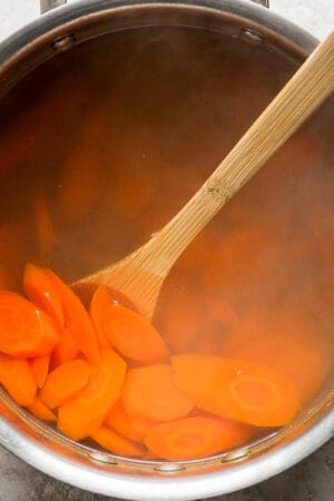 Top down shot of carrots being boiled in water in a saucepan with wooden spoon sticking out.