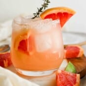 Straight on shot of a glass of a paloma cocktail with grapefruit wedges inside and on the rim with a sprig of thyme sticking out.