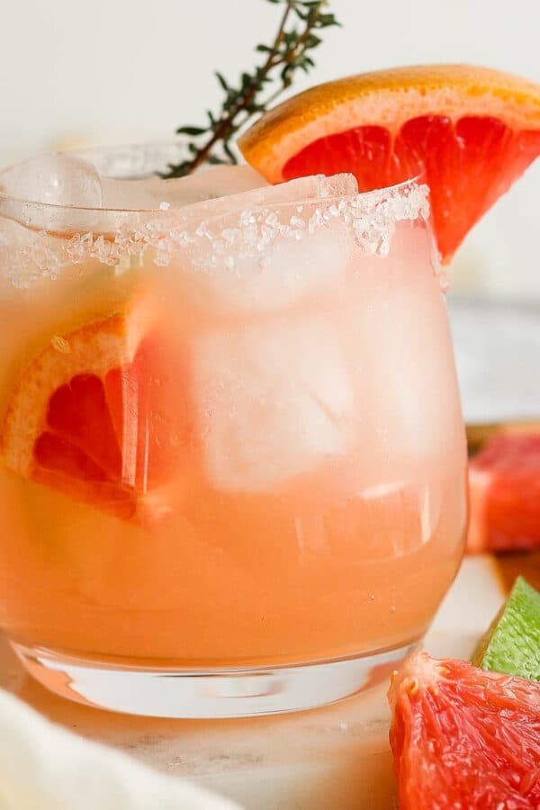 Straight on shot of a glass of a paloma cocktail with grapefruit wedges inside and on the rim with a sprig of thyme sticking out.
