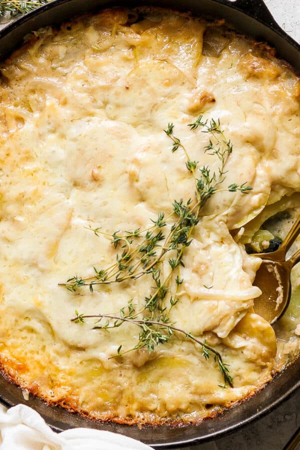 Top down shot of a cast iron skillet filled with potatoes au gratin gruyere with two spoons sticking out and fresh thyme sprigs on top.
