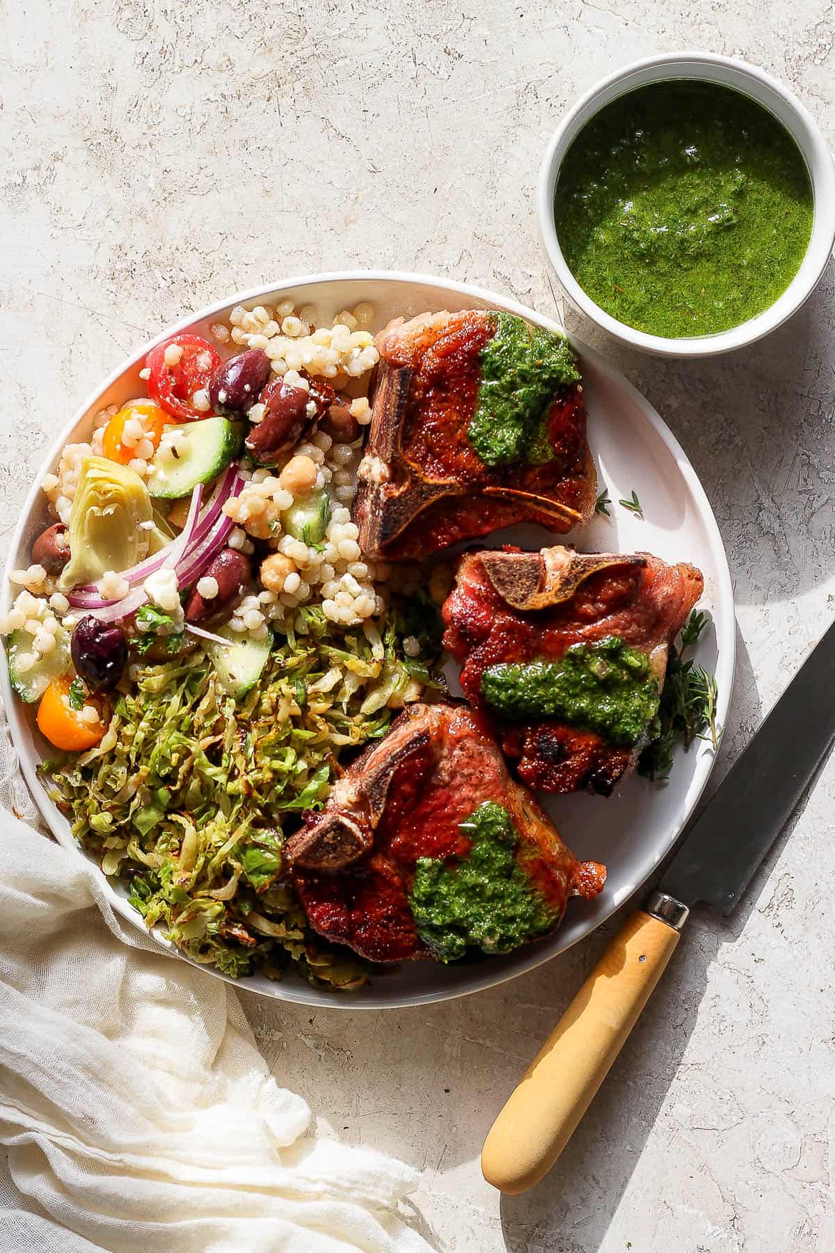 Lamb chops on a white plate with mint sauce on top and couscous salad and brussel sprouts on the side.