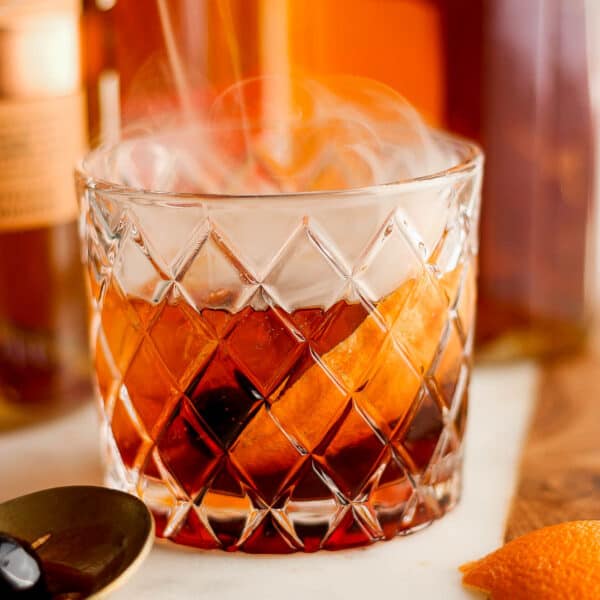 Straight on shot of a low ball glass with a smoked old fashioned inside with smoke coming out and a spoon next to it.