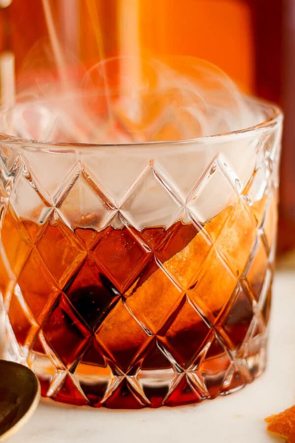 Straight on shot of a low ball glass with a smoked old fashioned inside with smoke coming out and a spoon next to it.