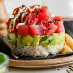 Straight on shot of a spicy tuna sushi stack with rice on the bottom, then cucumber and guacamole and then chunks of spicy tuna on a plate.