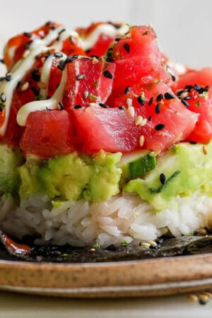 Straight on shot of a spicy tuna sushi stack with rice on the bottom, then cucumber and guacamole and then chunks of spicy tuna on a plate.