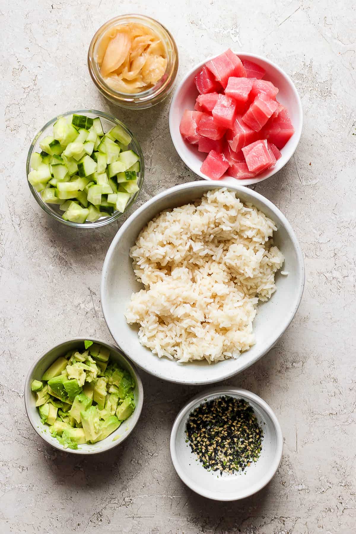 Ingredients for a sushi stack in small bowls with tuna chunks, chopped cucumber, rice, ginger and avocado. 