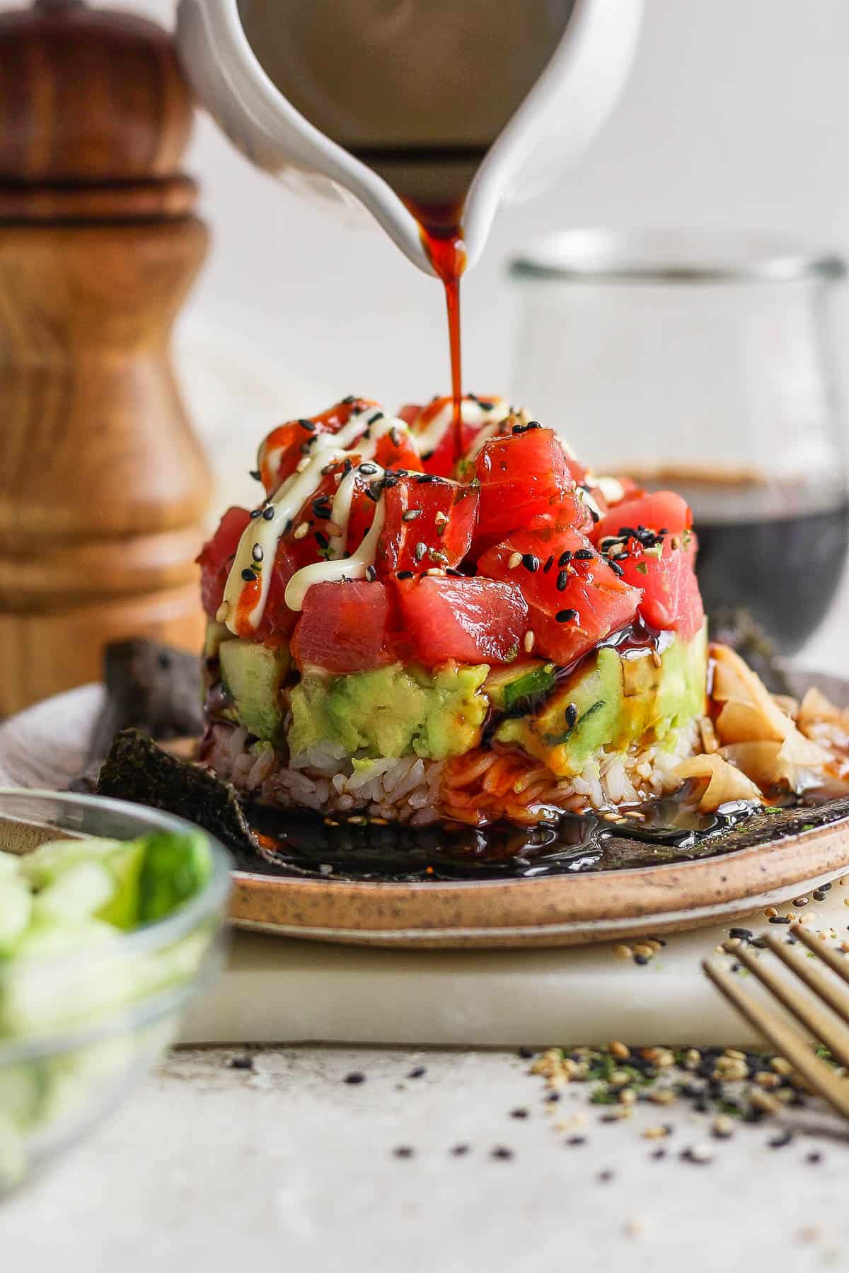 Straight on shot of a spicy tuna sushi stack with rice, avocado, cucumber and tuna layers with sesame seeds on top and someone pouring soy sauce on top. 