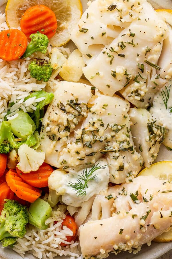 Top down shot of a plate with three pieces of baked cod with tartar sauce, rice and roasted vegetables.