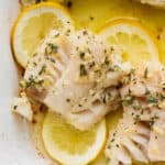 Top down shot of a piece of baked cod in a pan with three pieces of sliced lemon and melted butter around it.