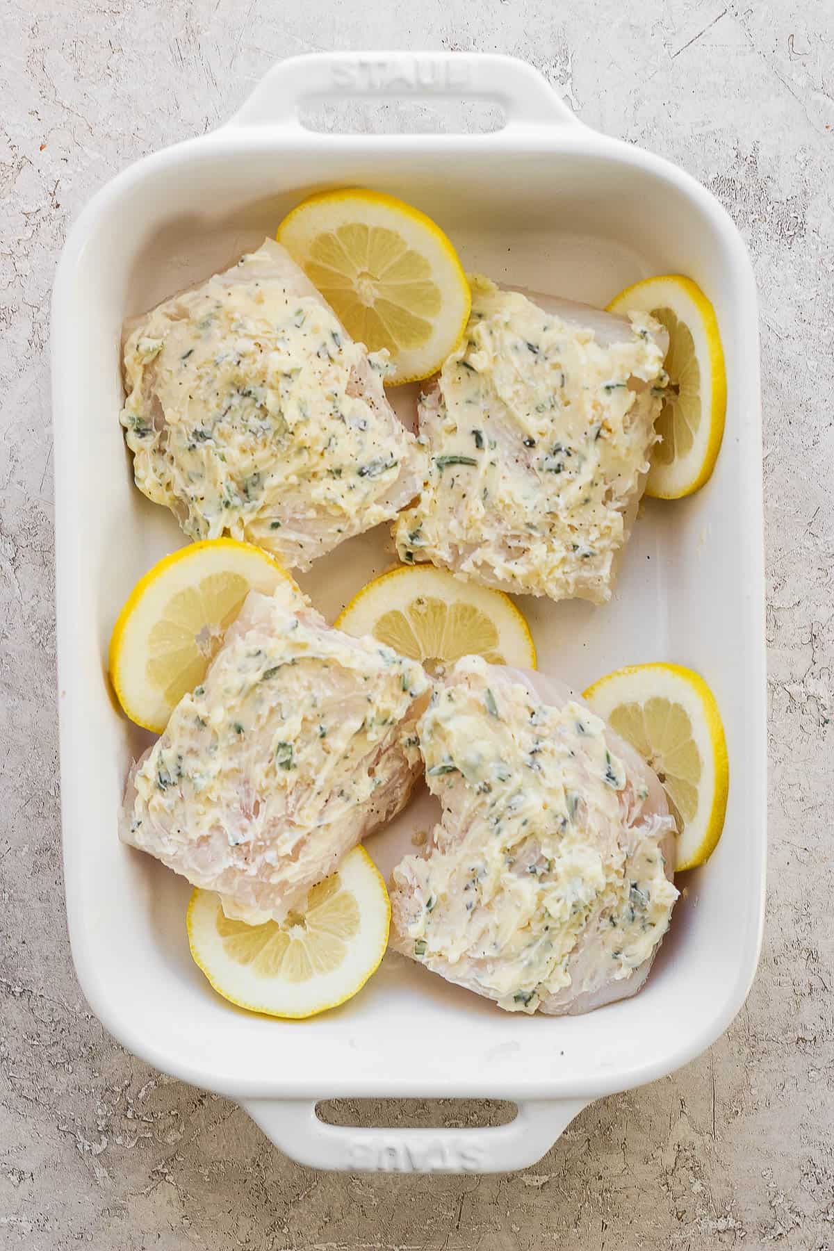 A white baking dish with cod fillets covered in herbed butter and surrounded by lemon slices.