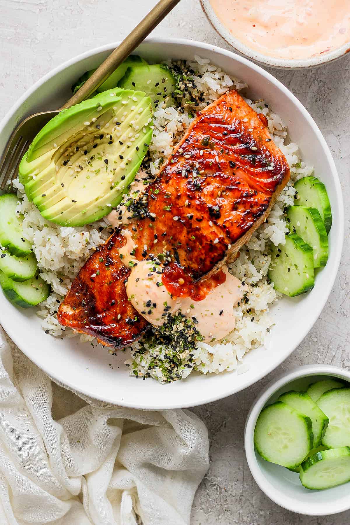 Baked salmon sushi bowl with salmon, cucumbers, sliced avocado, spicy mayo, and a salmon filet on top of rice. All garnished with furikake.