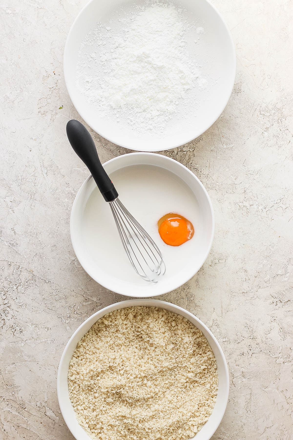 A bowl with corn starch next to a bowl with almond milk and an egg, next to a bowl with panko bread crumbs.
