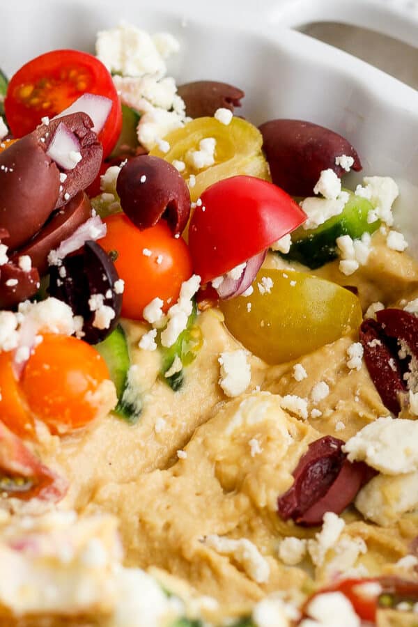 Side angle shot of a Greek layer dip in a dish with some of the bottom hummus layer exposed.