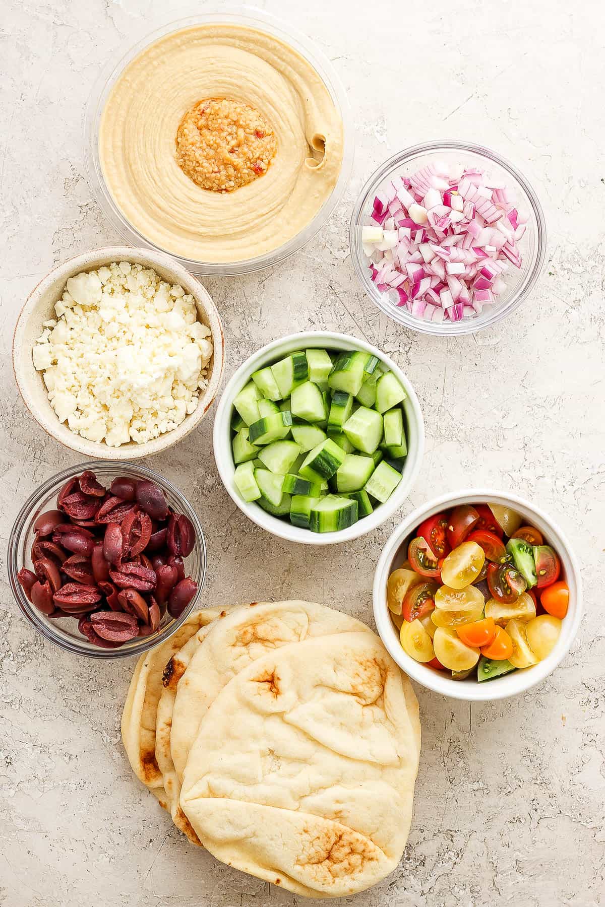 Individual containers of hummus, diced red onion, feta cheese, quartered and sliced cucumber, kalamata olives, halved cherry tomatoes, and naan bread.
