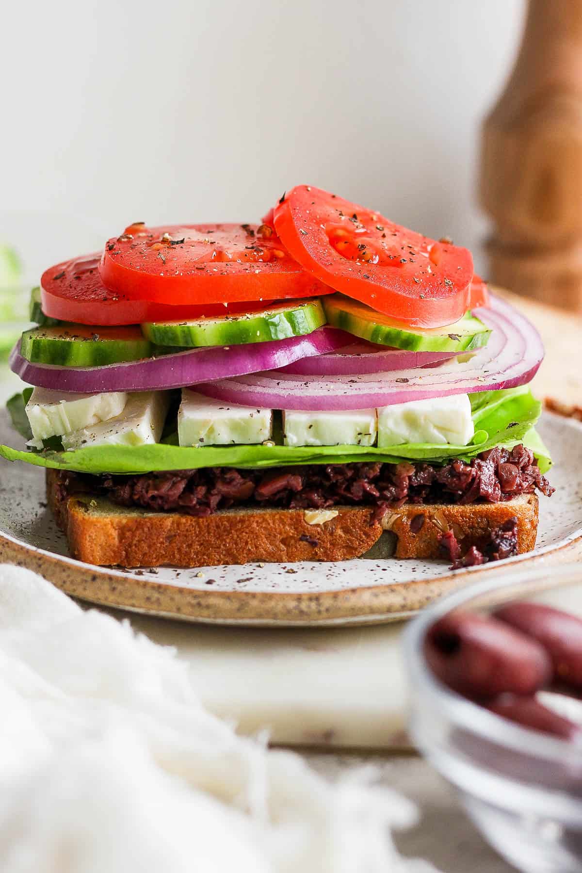 A piece of bread on a plate with the olive spread, lettuce, cheese, onions, cucumbers, tomato, salt and pepper on top.
