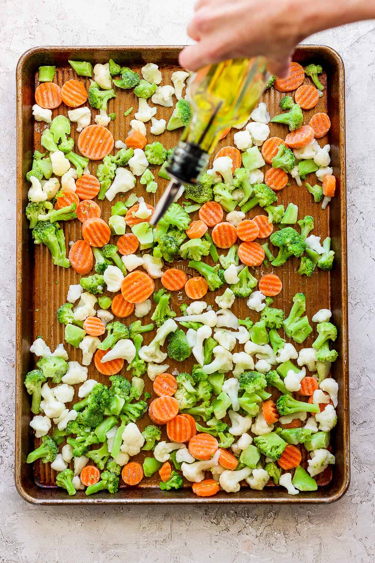 Frozen veggies on a rimmed baking sheet being drizzled with olive oil.