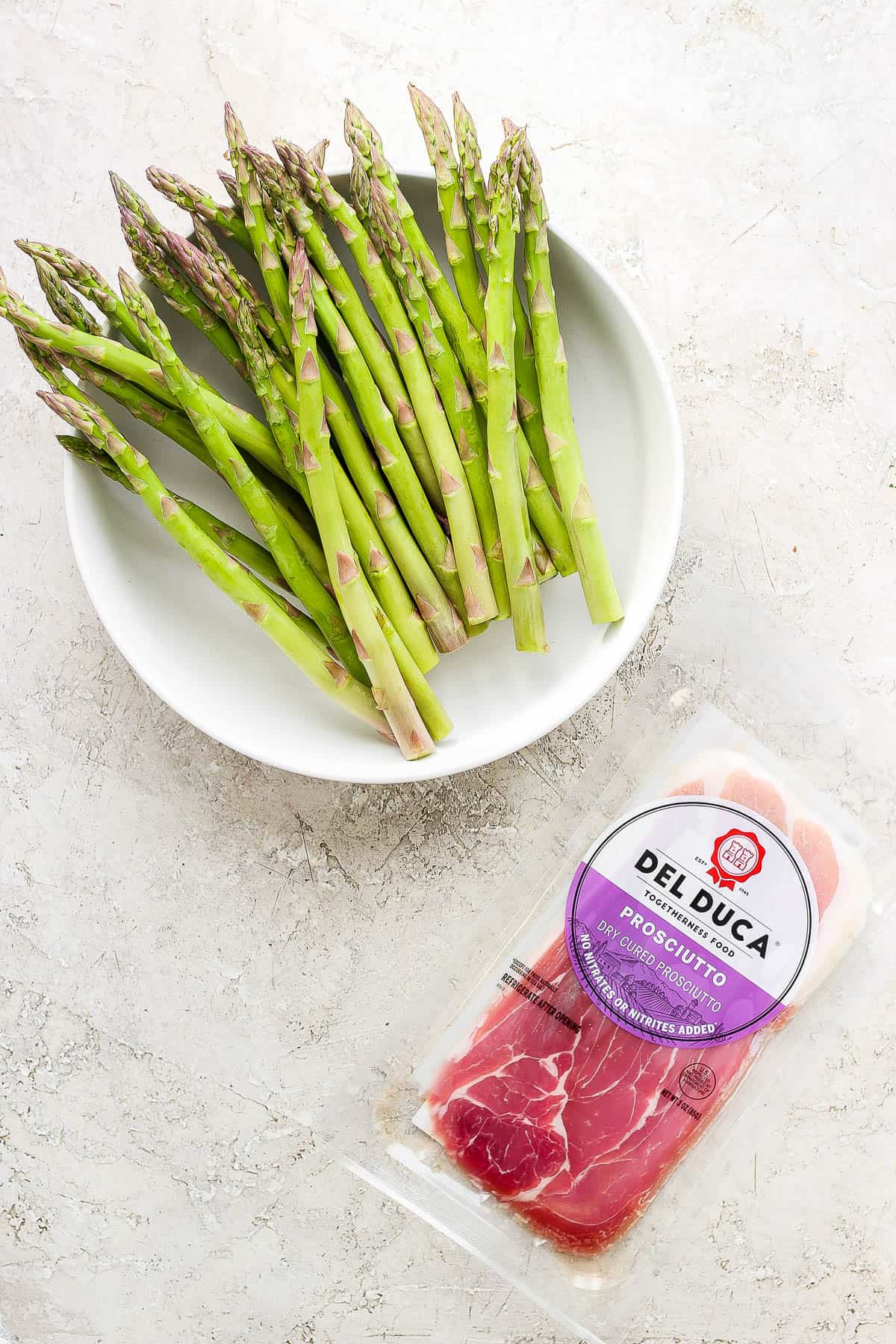 Trimmed asparagus in a white bowl next to a package of prosciutto.
