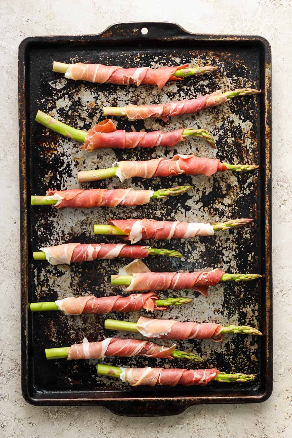 Several spears of prosciutto wrapped asparagus on a sheet pan before baking.
