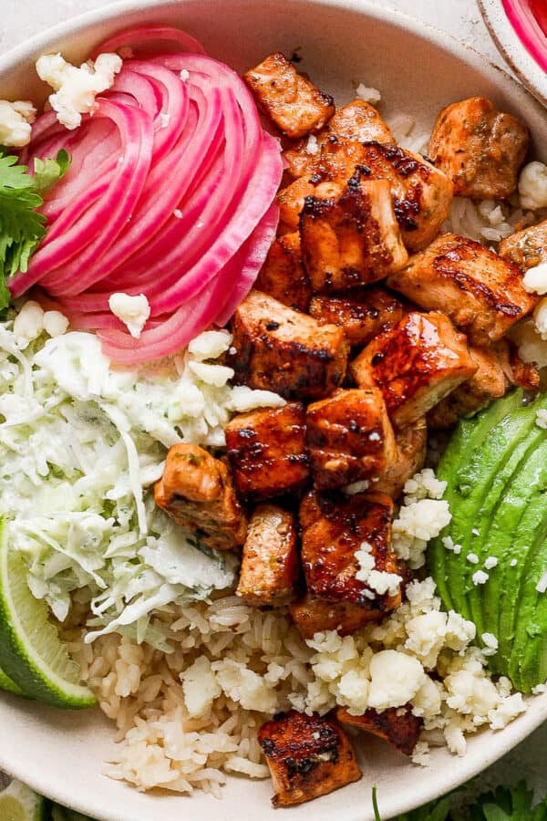 Top down shot of a salmon taco bowl with rice, limes, quick pickled onions, salmon pieces, avocado and slaw.