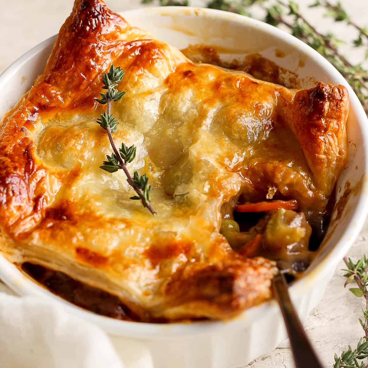 Side angle of a beef pot pie in a ramekin with puff pastry crust and a spoon sticking out.