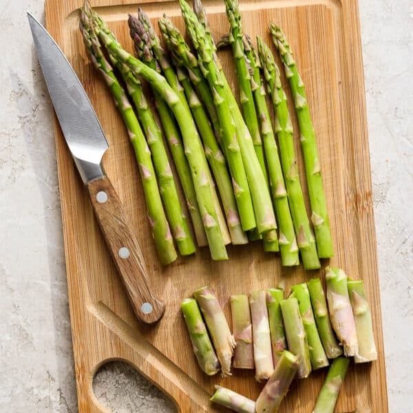 Top down shot of a cutting board with a bunch of asparagus on top with the ends trimmed and a knife next to them.