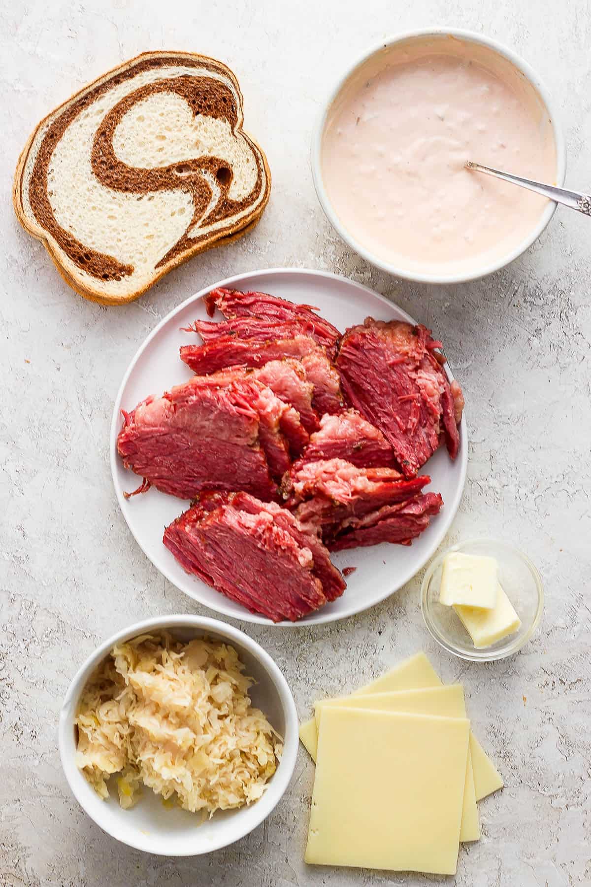 Two slices of rye bread, a bowl of thousand island dressing, a plate of corned beef, a bowl of butter, three slices of swiss cheese, and a bowl of saurkraut.