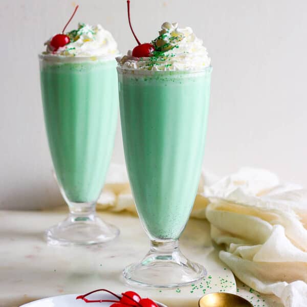 Straight on shot of two glasses filled with a shamrock shake and topped with whipped cream, a cherry and green sprinkles.