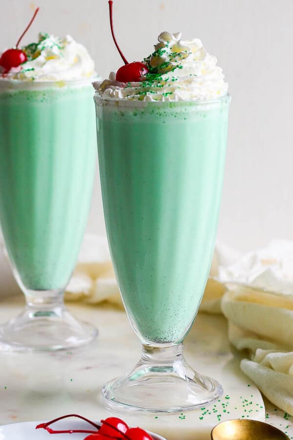 Straight on shot of two glasses filled with a shamrock shake and topped with whipped cream, a cherry and green sprinkles.