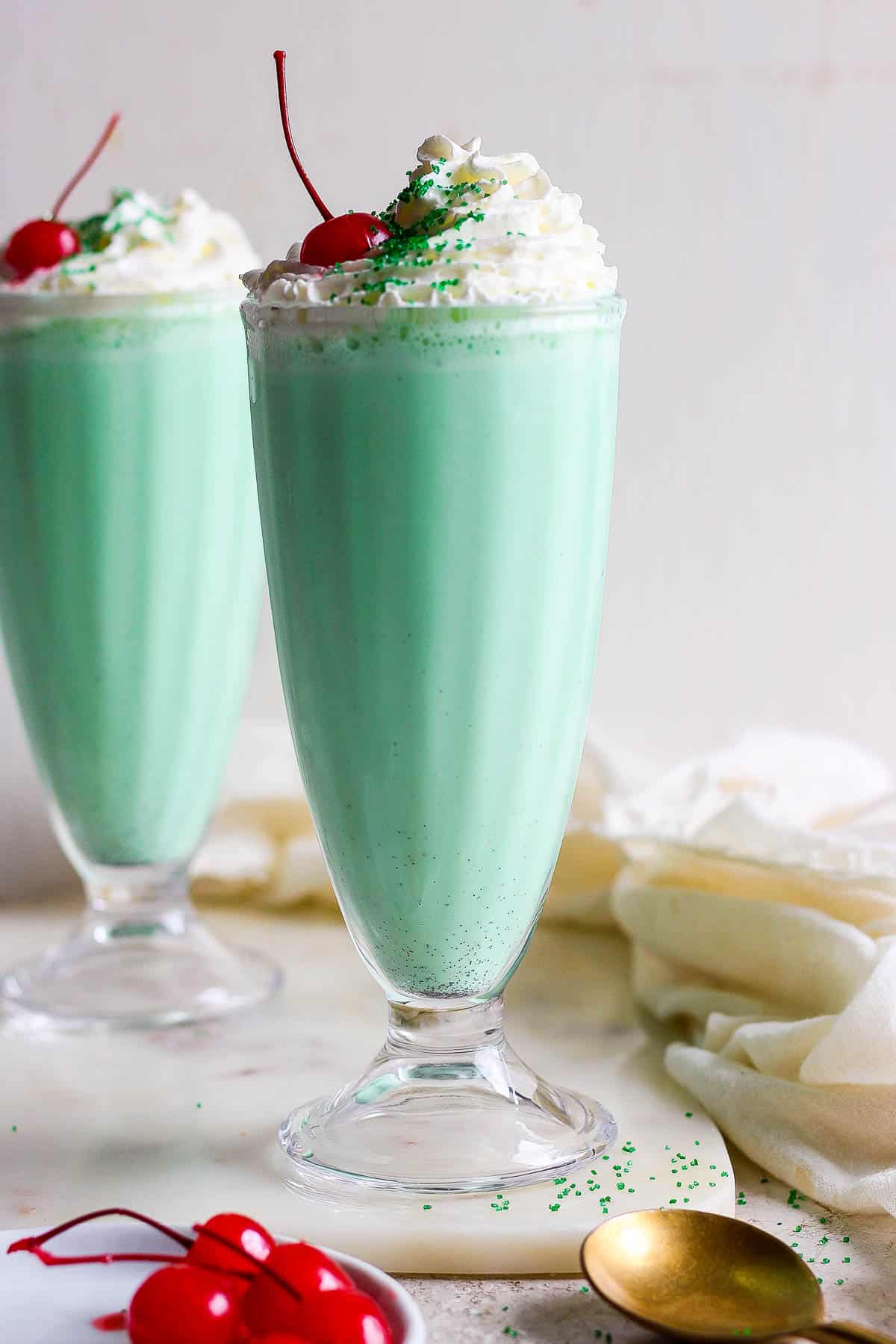 Two glasses filled with the shamrock shake mixture and topped with whipped cream, a cherry, and green sprinkles.