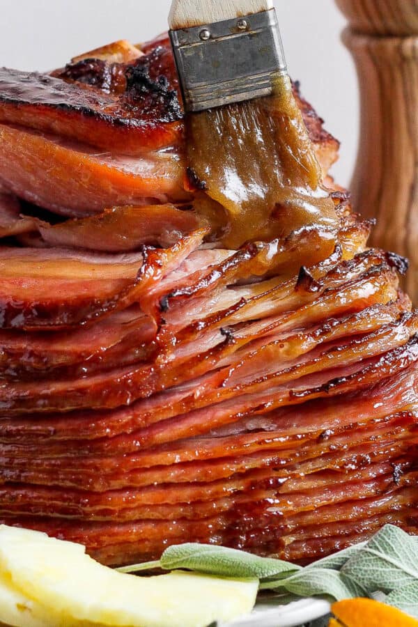A side shot of a spiral ham with someone brushing a 3 ingredient glaze over it with a small brush.