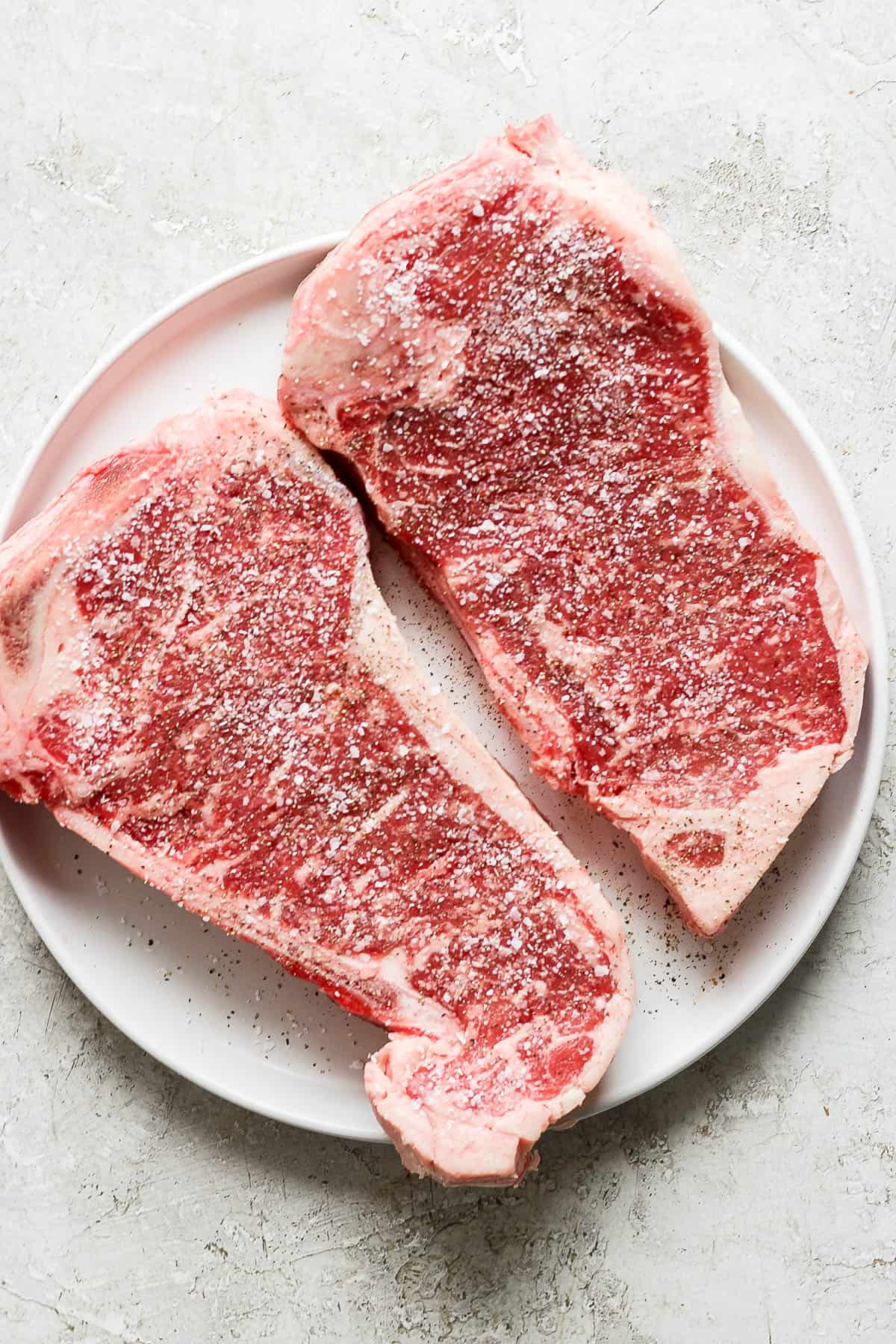Raw steaks seasoned with salt and pepper on a white plate.