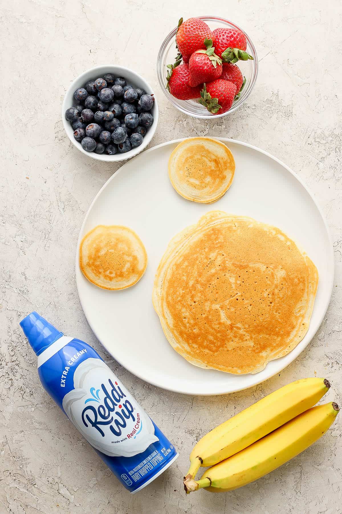 Three pancakes on a plate surrounded by fresh bananas, a can of whipped cream and bowls of berries.