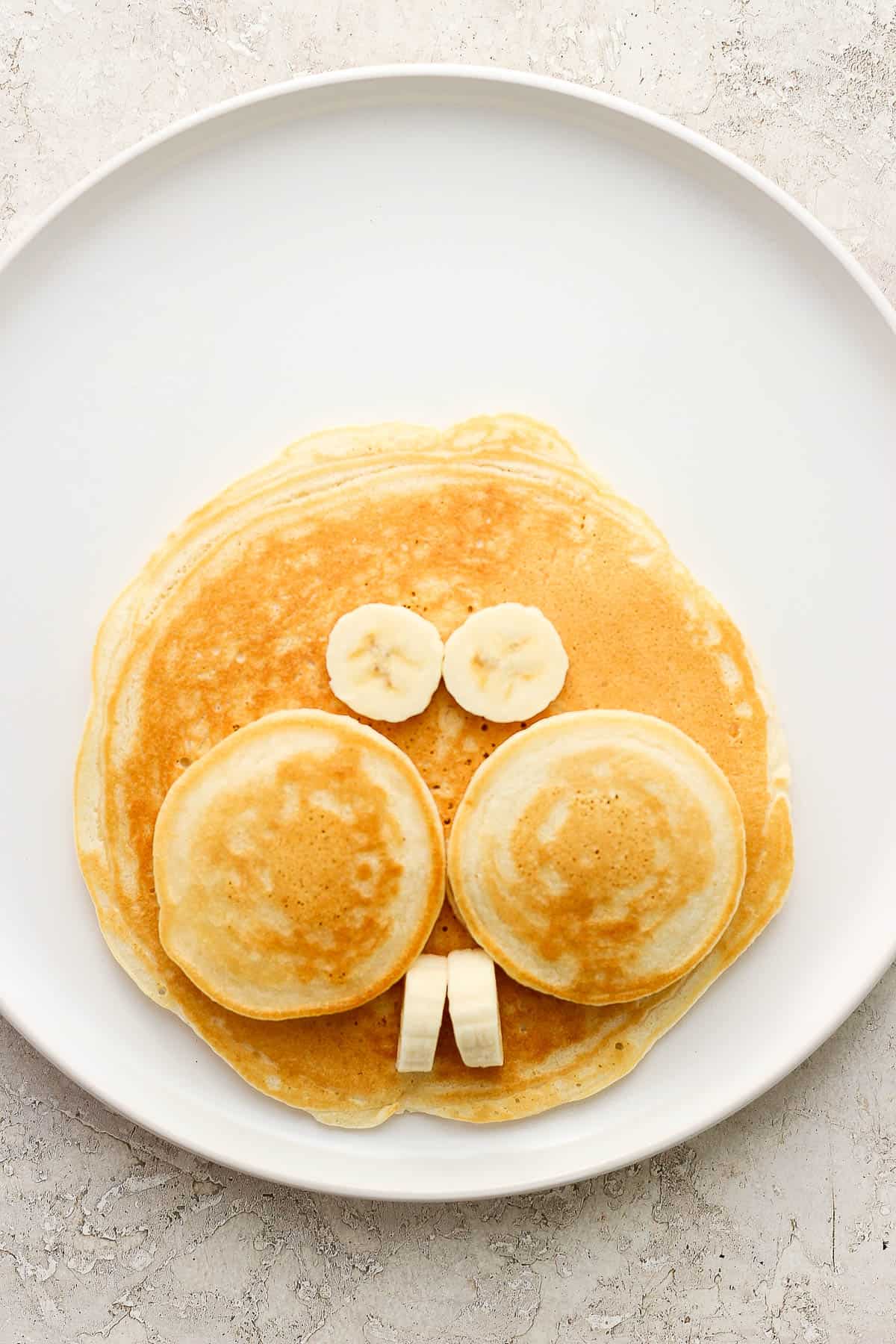 One large pancake with two small pancakes as cheeks and banana slices as eyes and teeth.