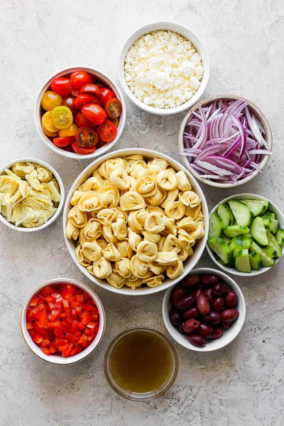 Individual bowls of feta cheese, sliced cherry tomatoes, sliced red onion, artichoke hearts, cheese tortellini, sliced cucumbers, olives, diced red bell pepper, and salad dressing. 