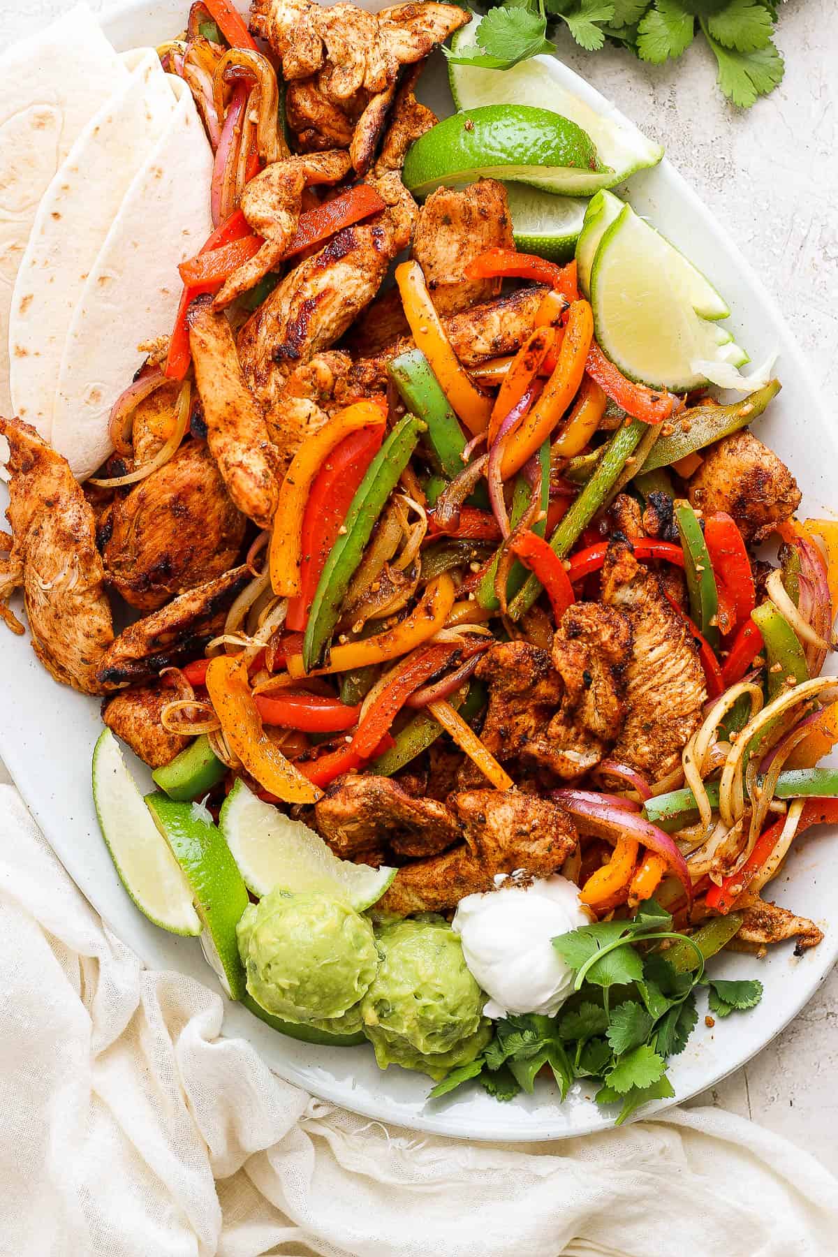 A serving platter of grilled chicken fajitas with tortillas, guacamole, sour cream, lime wedges, and cilantro.