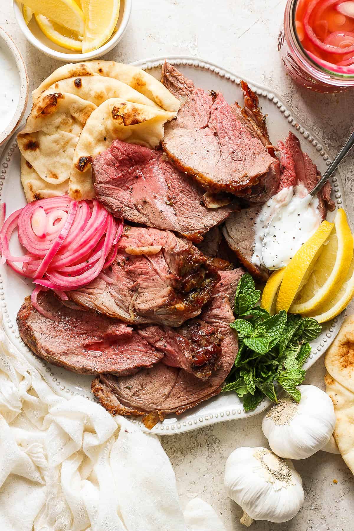 Slices of grilled leg of lamb on a plate with fresh mint, lamb sauce, pickled red onions, naan bread, etc.