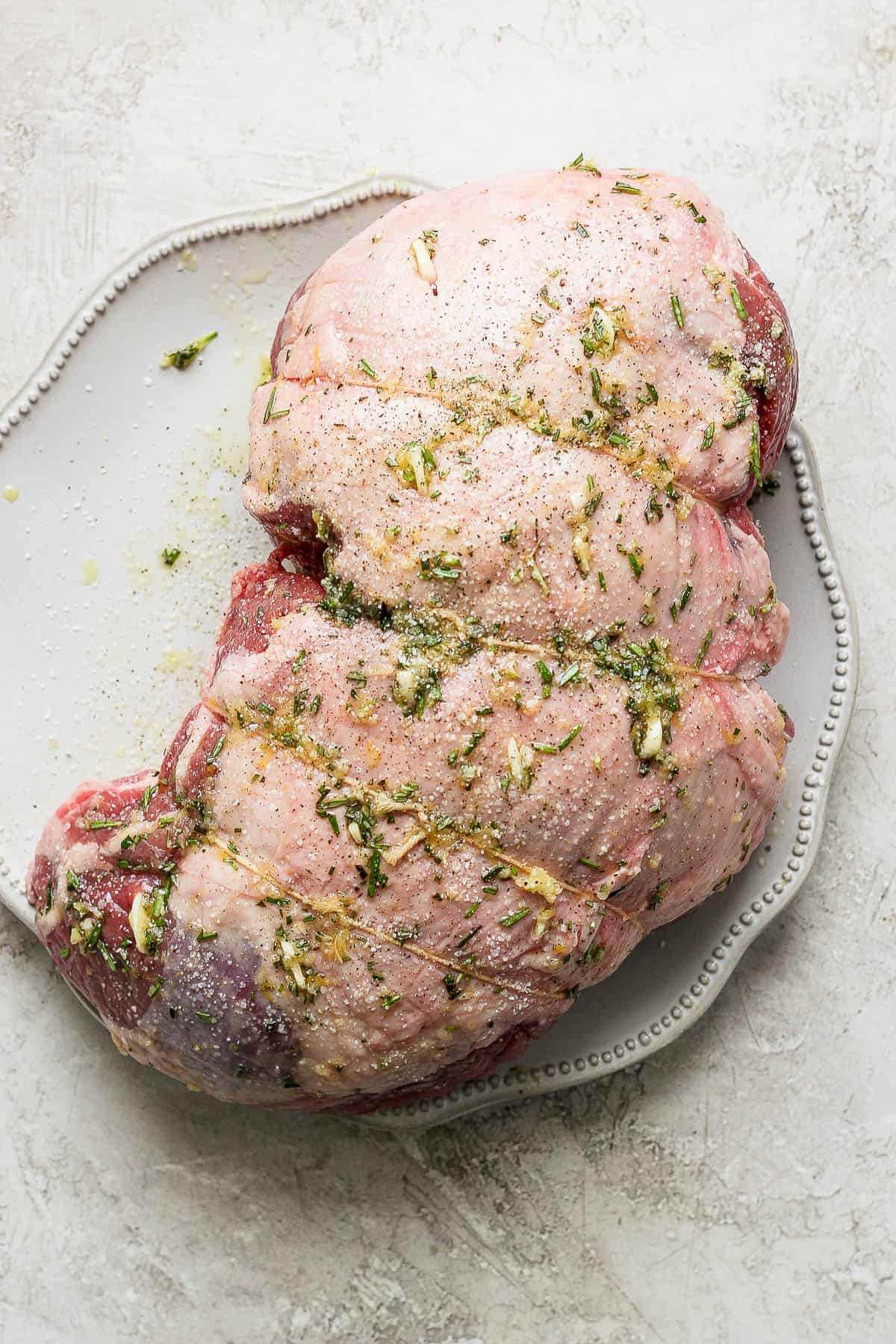 Boneless leg of lamb on a plate with marinate on the outside and slices of garlic in it.