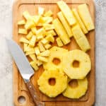 Top down shot of a cutting board with a knife, pineapple chunks, circles and spears.