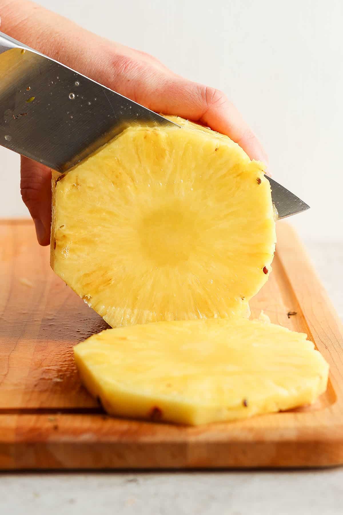 A whole pineapple on it's side with the end, top, and skin cut off being cut into rounds.