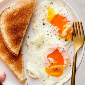 Someone holding a plate with two over easy eggs, a gold fork and a piece of toast.
