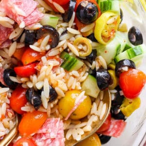 Top down shot of bowl of orzo pasta salad with olives, tomatoes, cucumbers and salami with two spoons sticking out.