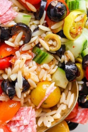 Top down shot of bowl of orzo pasta salad with olives, tomatoes, cucumbers and salami with two spoons sticking out.
