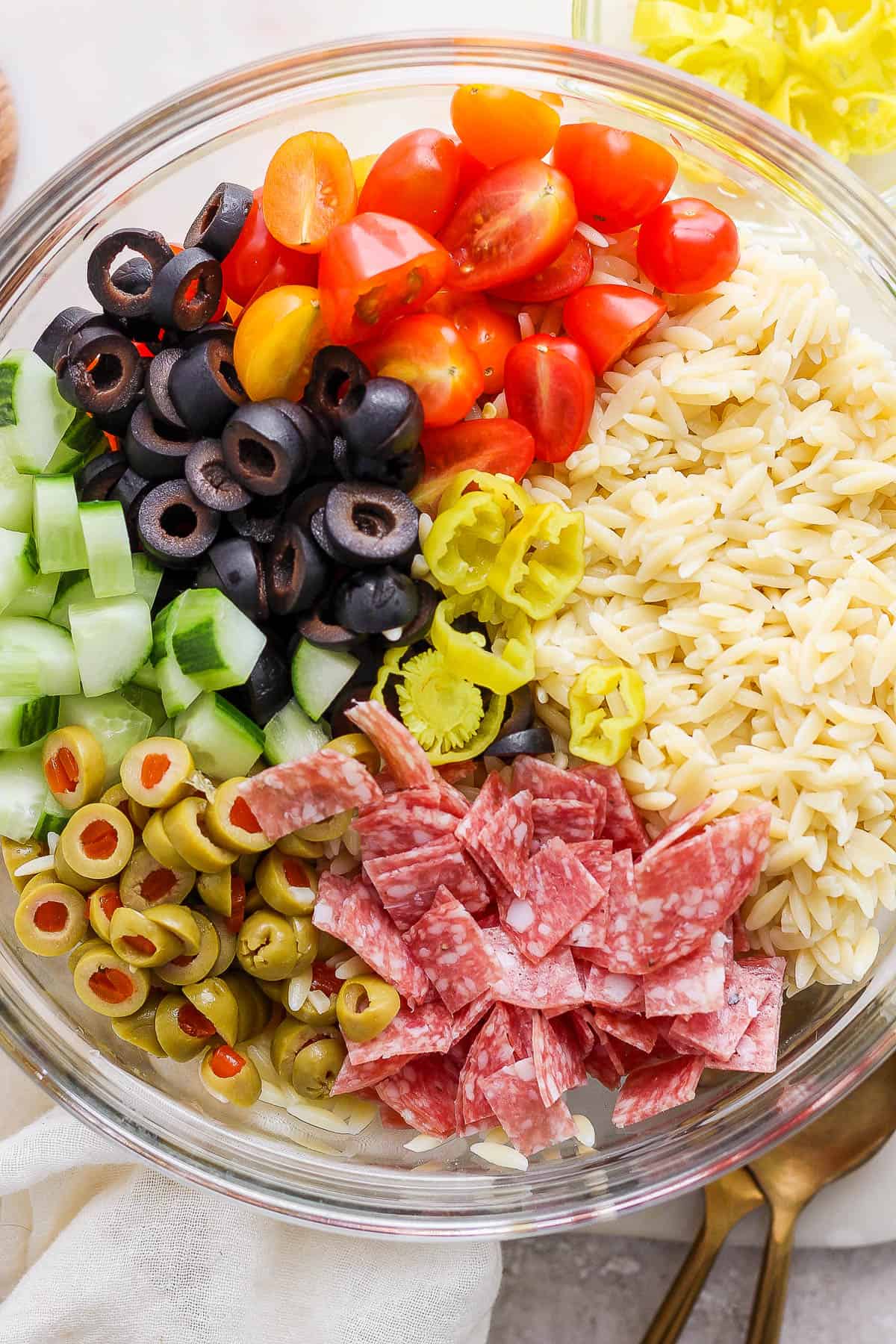 Cooked orzo, sliced cherry tomatoes, black and green olives, cucumbers, salami, and pepperochini in a large mixing bowl.