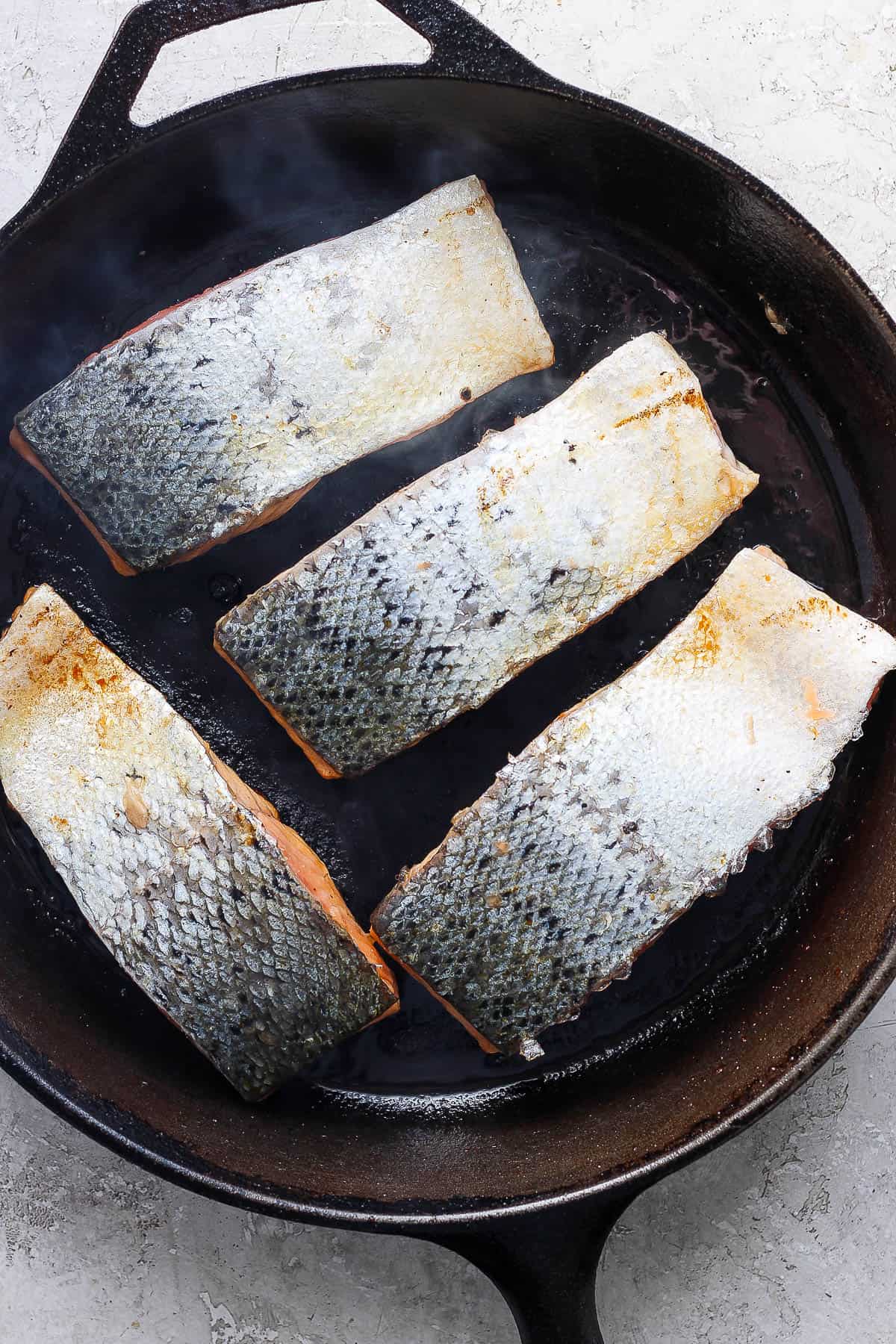 Salmon fillets searing in a cast iron skillet with the skin-side up.