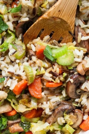 Top down shot of a wooden spoon sticking out of a pan of wild rice pilaf with carrots, celery and chopped parsley.