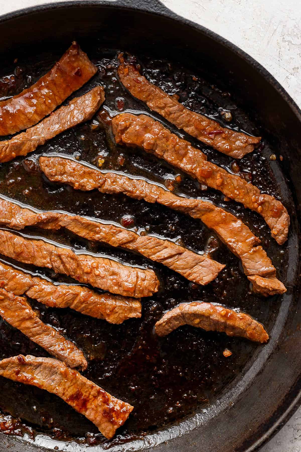 Strips of marinated beef being seared in a cast iron skillet.