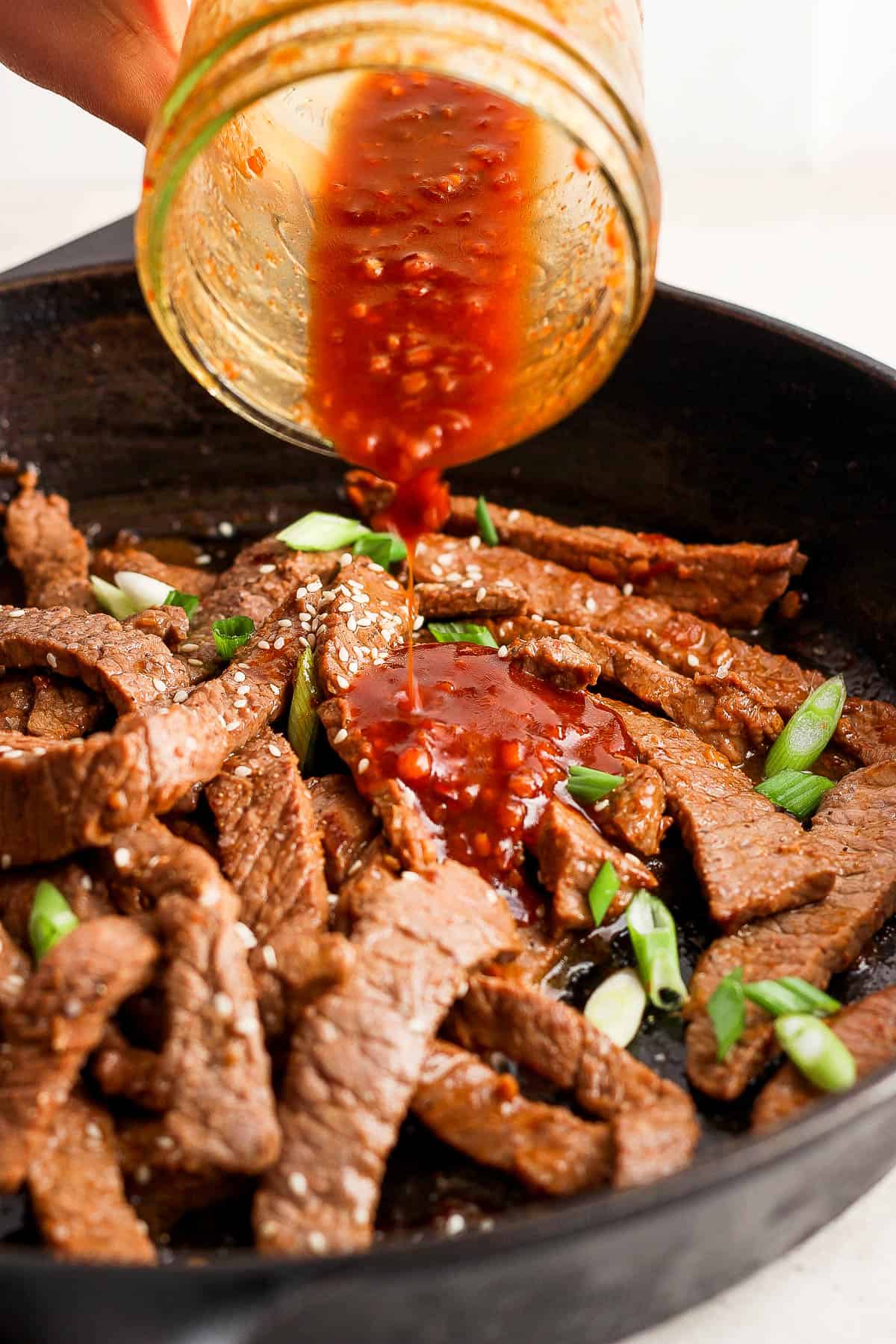 Extra sauce being poured over the beef strips in the skillet topped with sesame seeds and sliced green onions.