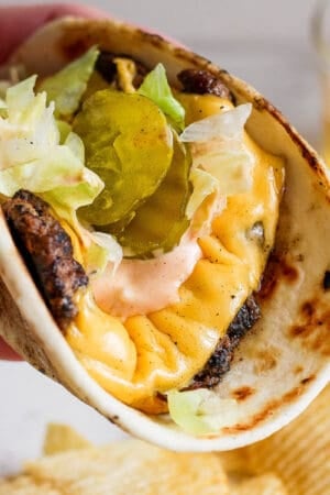 Someone holding a smash burger taco with shredded lettuce, burger sauce and pickles.